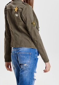 Chaqueta de mezclilla verde oliva con un diseño ajustado, que presenta dos parches dorados en forma de estrella y cierre de botones, combinada con jeans azules desgastados.