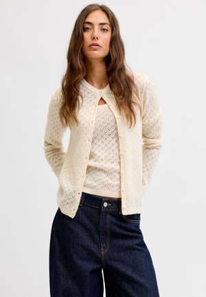 Femme aux longs cheveux bruns porte un cardigan crème boutonné sur un top en maille assorti et un jean taille haute bleu foncé sur un fond uni.