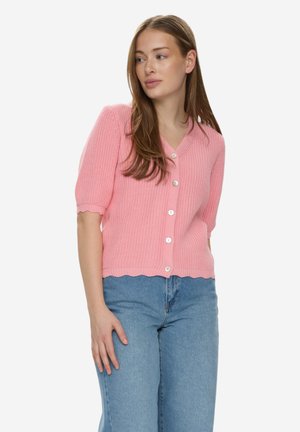 Mujer que lleva un cárdigan de punto de manga corta color rosa con botones y vaqueros azul claro, mirando hacia su derecha con expresión neutral.