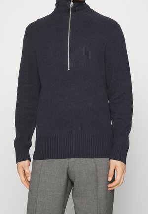 Homme portant un pull en maille côtelée bleu marine foncé avec un col à demi-fermeture éclair et un pantalon gris taillé sur fond blanc.
