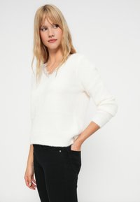 Nuori nainen, jolla on pitkä vaalea hiukset, pukeutuu valkoiseen pörröiseen neuleeseen pitsireunaisella paidan päällä ja mustiin housuihin, käsi taskussa, katsoo sivulle.