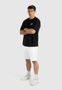 Schwarzes, übergroßes T-Shirt mit rundem Halsausschnitt und weißem Logo, kombiniert mit weißen Shorts und weißen Sneakers. Einheitliche Farbe, lässiges Design.