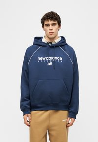 Jonge man met donker krullend haar, gekleed in een marineblauwe New Balance hoodie en beige New Balance trainingsbroek, staande tegen een effen achtergrond.