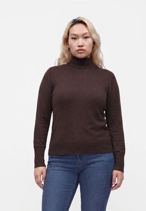 Personne aux longs cheveux blonds portant un pull à col roulé marron foncé et un jean bleu, debout devant un fond clair uni.