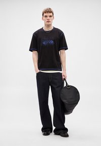 T-shirt preta com detalhes azuis e gráfico "HUGO", combinada com calças de ganga escuras. Modelo segura uma mala redonda preta. Texturas suaves.