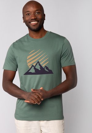 Lächelnder Mann mit dunkler Hautfarbe, der ein grünes T-Shirt mit Bergmotiv und beige Shorts trägt, steht vor einem schlichten hellen Hintergrund.