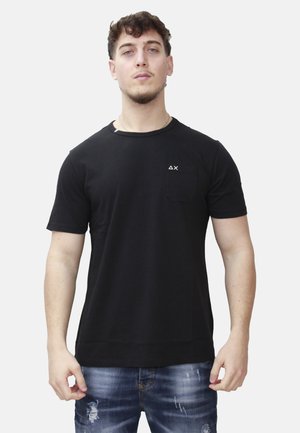 Uomo con capelli corti e ricci che indossa una T-shirt nera con un piccolo logo bianco a forma di triangolo e X sul petto, abbinata a jeans blu strappati.