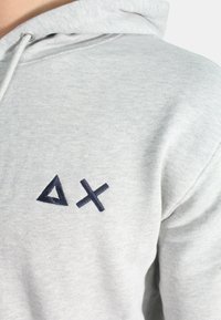 Felpa grigia realizzata in un tessuto morbido, con ricami geometrici in blu scuro a forma di triangolo e croce sulla parte sinistra del petto.