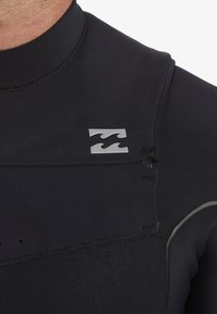 Primo piano di un colletto di una muta nera con un piccolo logo a onda argentato sulla spalla e dettagli di cucitura visibili.