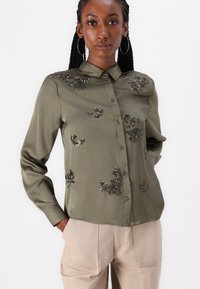 Olive zaļas satīna krekliņš ar apkakli un priekšējiem pogām, greznoti ar melniem ziedu fliteriem krūšu un piedurkņu rajonā.