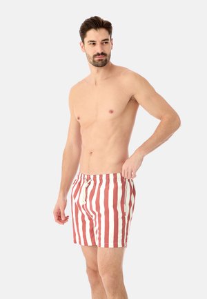 Homme torse nu avec une barbe portant un short rayé vertical rouge et blanc, ajustant la taille, debout devant un fond clair uni.