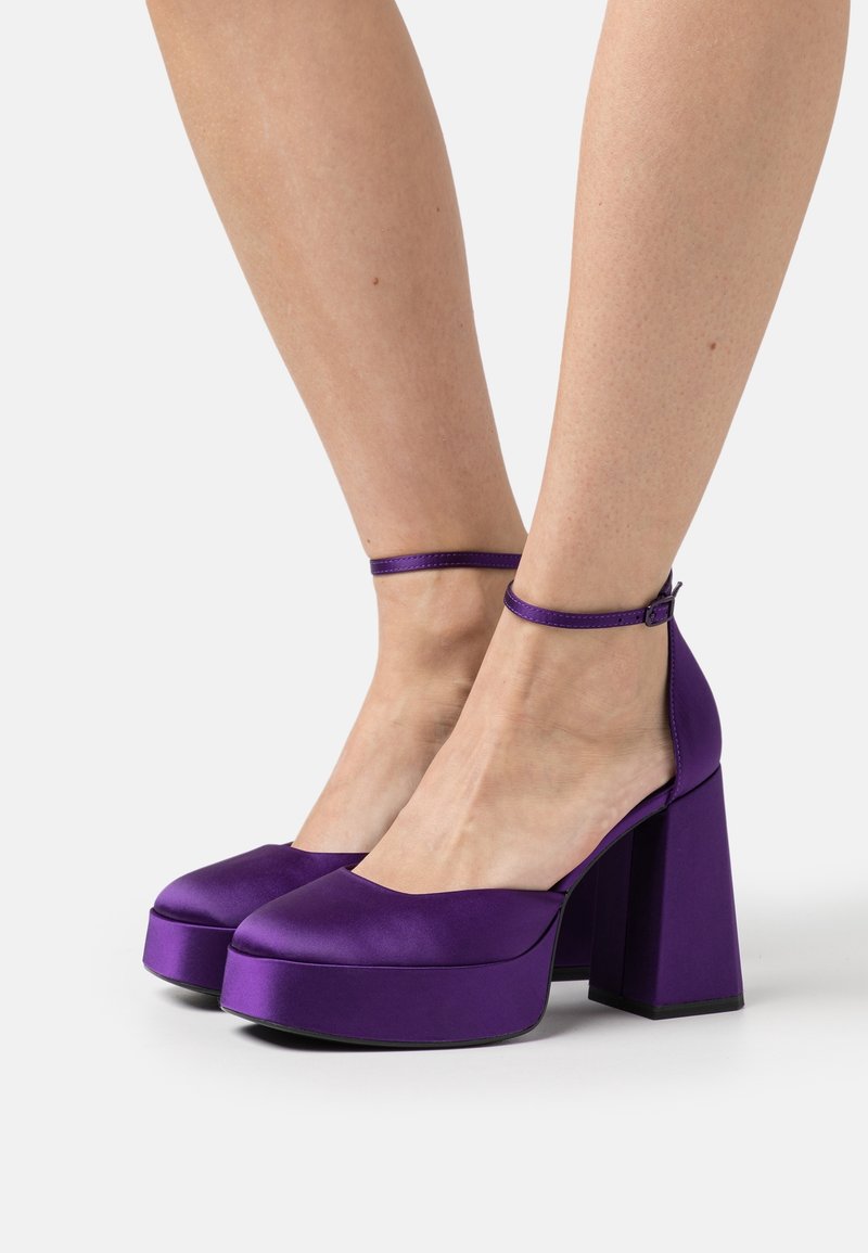 Jambes portant des escarpins plateforme en satin violet avec brides à la cheville et talons blocs hauts et épais, debout sur une surface blanche.