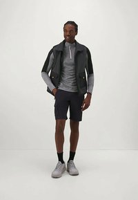 Svart vest over en grå langermet topp med glidelås, kombinert med marineblå shorts og grå sneakers. Minimalistisk design med praktiske lommer.