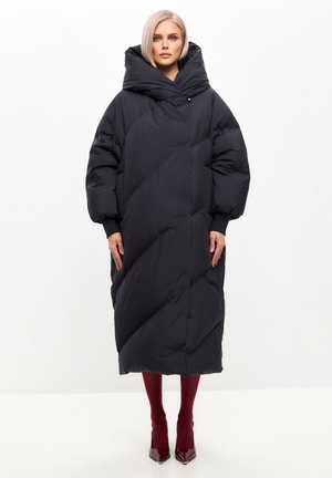Dikke zwarte mantel tot op de knie met een oversized capuchon, diagonale stiksels en volumineuze mouwen. Gemaakt over bordeauxrode panty's en hakken.