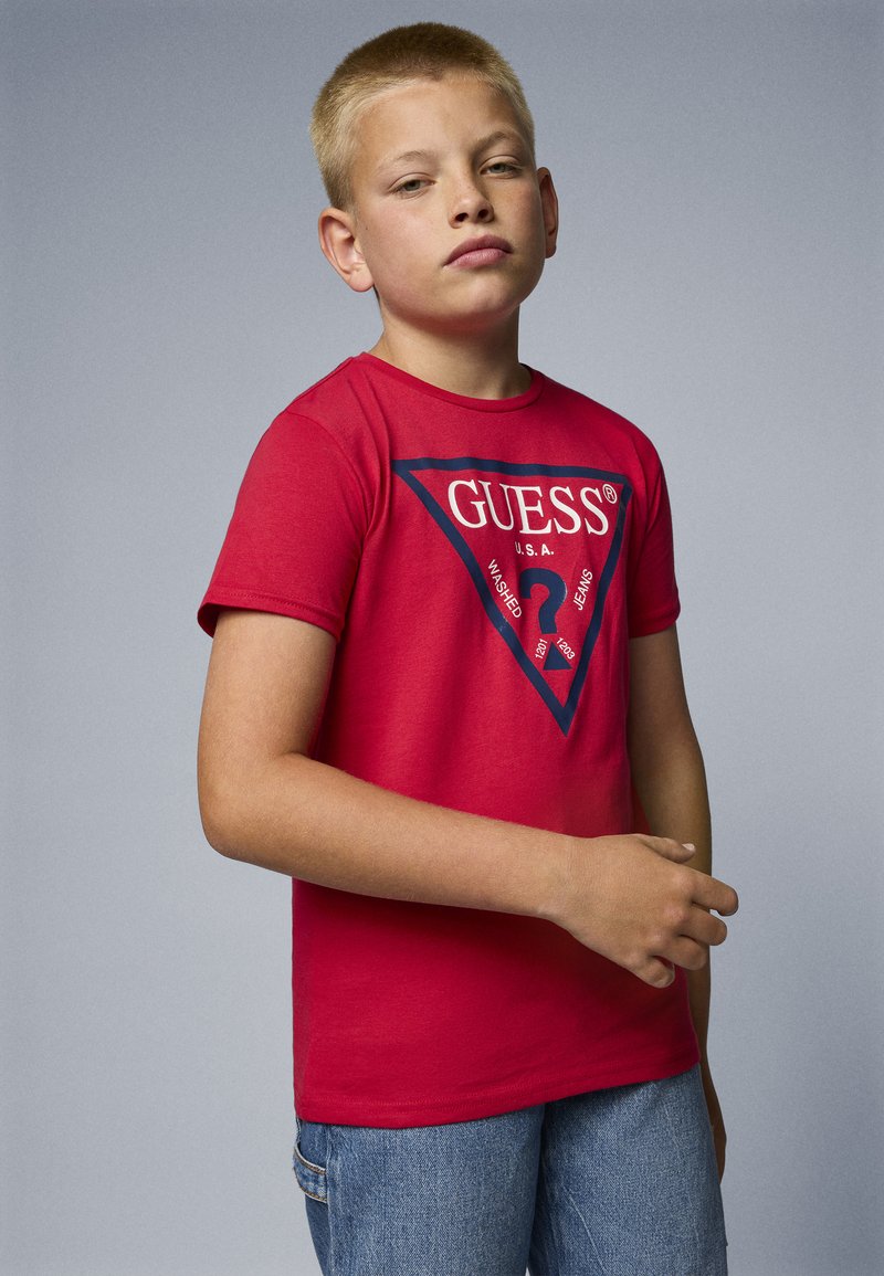 Rød bomull t-skjorte med korte ermer og en stor marineblå trekantlogo med "GUESS" i hvitt, sammen med blå denimshorts.