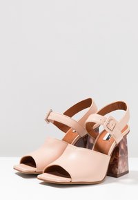 Béžové kožené sandály na podpatku, otevřené na špičce, s řemínky kolem kotníku a čtvercovými blokovými podpatky s želvovinovým vzorem na bílém povrchu.