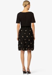 Robe noire avec un haut ajusté et des manches courtes, présentant une jupe en dentelle florale avec des accents dorés et un ourlet au genou. Fermeture éclair au dos.