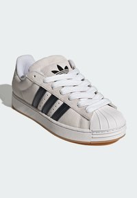 Svetlosivá semišová teniska s bielymi šnúrkami a gumovou špičkou. Má čierne tri pruhy a logo na jazyku. Podrážka z gumy farby gumy.