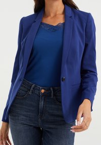 Blå blazer med slag, enkel knäppning och två framfickor, bärs över en blå linne med spetsdetaljer, tillsammans med mörka jeans.