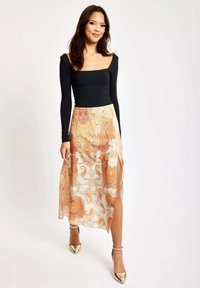 Black long-sleeve top paired with a sheer cream skirt featuring orange paisley patterns and side slits; gold heeled sandals complete the look.