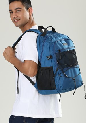 Man draagt een wit T-shirt en heeft een grote blauwe rugzak met meerdere ritsvakken en gaas-zijvakken op de rug.