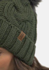 Green knitted beanie with a textured pattern. Features a brown leather label. Topped with a black pom-pom. Soft, warm material.