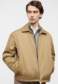 Στενή ανοιχτό καφέ ζακέτα με φερμουάρ, με υφή στην επιφάνεια, γιακά που διπλώνει προς τα κάτω και ελαστικές μανσέτες, φορεμένη πάνω από ένα γκρι ζακέτα με φερμουάρ και επίπεδα στρώματα.