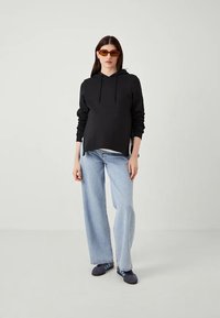 Woman wearing black hoodie, light blue wide-leg jeans, navy sneakers with blue stripes, and orange-tinted sunglasses standing against plain background.