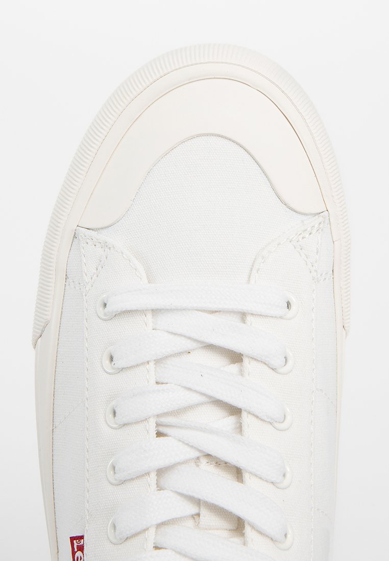 Zapatilla de lona blanca con punta redonda, cordones planos blancos, lados texturizados y suela de goma con borde estriado. Detalle del logotipo rojo pequeño.