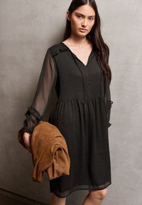 Vestido negro con estampado, escote en V, mangas transparentes y detalles de volantes abullonados, acompañado de una chaqueta de ante marrón plegada.