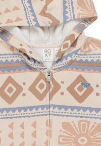 Felpa beige con motivo tribale in marrone e blu. Presenta una zip frontale, un interno morbido e un logo a forma di cuore sul petto.