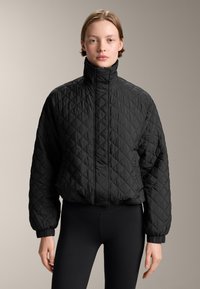 Giacca trapuntata nera con colletto alto, polsini e orlo elasticizzato. Tessuto leggero con superficie texture. Abbinata a leggings neri.