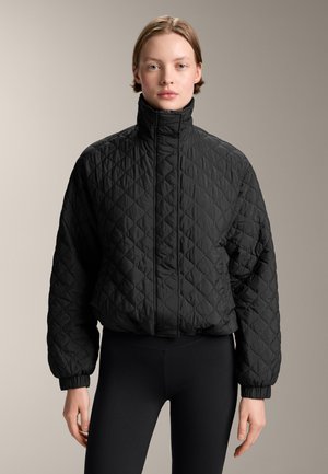 Gequilted zwarte jas met hoge kraag, elastische manchetten en zoom. Lichtgewicht stof met een gestructureerd oppervlak. Gecombineerd met zwarte leggings.