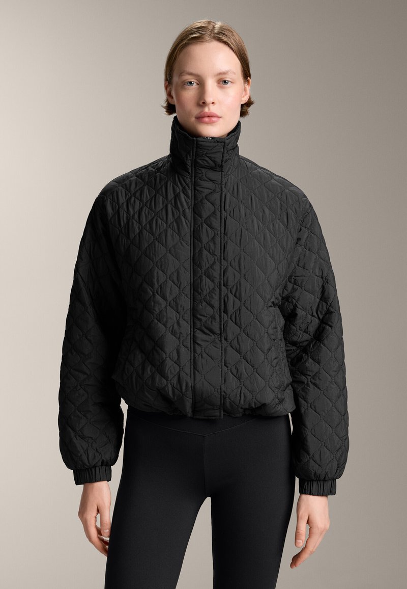 Giacca trapuntata nera con colletto alto, polsini e orlo elasticizzato. Tessuto leggero con superficie texture. Abbinata a leggings neri.