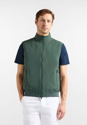 Gilet verde senza maniche in tessuto liscio, caratterizzato da un colletto alto, cerniera frontale e tasche laterali, indossato sopra una camicia blu navy e pantaloni bianchi.