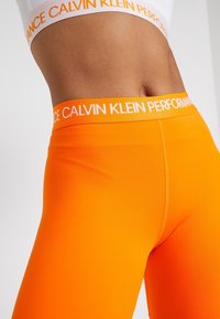Jasnopomarańczowe legginsy wykonane z elastycznego materiału, z szerokim pasem w talii z białym napisem "CALVIN KLEIN PERFORMANCE".
