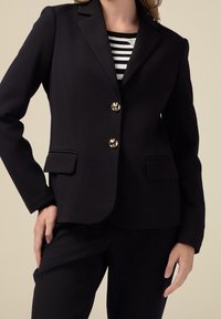 Blazer noir sur mesure avec une fermeture à trois boutons, des boutons en métal doré, des épaules structurées et des poches avant ; texture lisse et coupe ajustée.