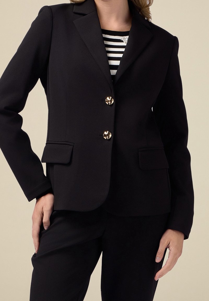 Blazer noir sur mesure avec une fermeture à trois boutons, des boutons en métal doré, des épaules structurées et des poches avant ; texture lisse et coupe ajustée.