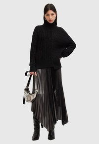 Pulover negru tricotat cu model împletit, combinat cu o fustă plisată neagră și cizme negre până la genunchi. Ține o geantă metalică argintie.