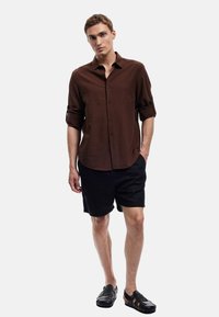 Bruine blouse met opgerolde mouwen, gecombineerd met zwarte shorts en zwarte sandalen. Model poseert tegen een eenvoudige witte achtergrond.