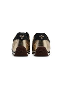 Ett par guld- och svartsneakers sedda bakifrån, vilket visar bruna strukturerade sulor och en bi-logotyp på klacken.