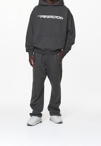 Grå huvtröja med "PEGADOR"-logotyp i vitt, tillsammans med matchande grå sweatpants. Båda är gjorda av mjukt tyg med en avslappnad passform. Vita sneakers.