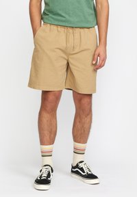 Beige katoenen shorts met een elastische tailleband en trekkoord, gedragen met zwarte sneakers en crème sokken met kleurrijke strepen.