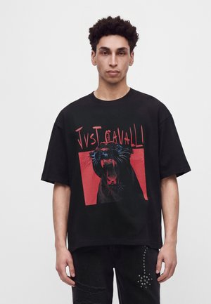 Νέος άνδρας φοράει μαύρο φαρδύ T-shirt με έναν βρυχόμενο μαύρο πάνθηρα και κόκκινο κείμενο "JVST CAVALLI" σε κόκκινο τετράγωνο φόντο.