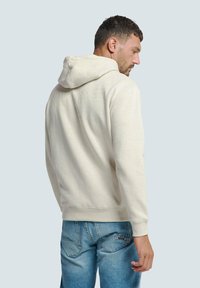 Licht beige hoodie met een trekkoordcapuchon, ribgebreide boorden en zoom, met minimale reliëfbranding op de achterkant. Gepaard met blauwe denim jeans.