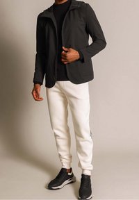 Czarna kurtka z zamkiem błyskawicznym i kieszeniami, czarna koszula, białe spodnie dresowe z tekstem po bokach oraz czarne sneakersy z kontrastową białą podeszwą.