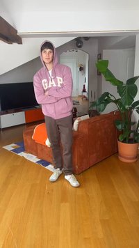 Mladi moški v roza kapucarju GAP in rjavih hlačah stoji z rokami prekrižanimi v dnevni sobi z oranžnim kavčem in veliko sobno rastlino v loncu.