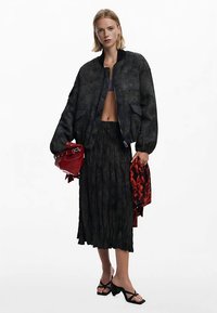 Veste bomber noire à finition texturée, jupe plissée noire, sandales noires et sac à main rouge. Les accessoires comprennent une écharpe rouge à motif.