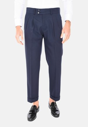 Pantaloni pieghettati blu navy con una texture liscia, dotati di due bottoni frontali e risvolti; abbinati a mocassini neri.