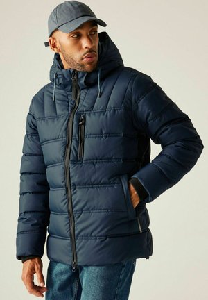 Mężczyzna w granatowej pikowanej kurtce z kapturem i szarej czapce, patrzący w bok, z jedną ręką w kieszeni.